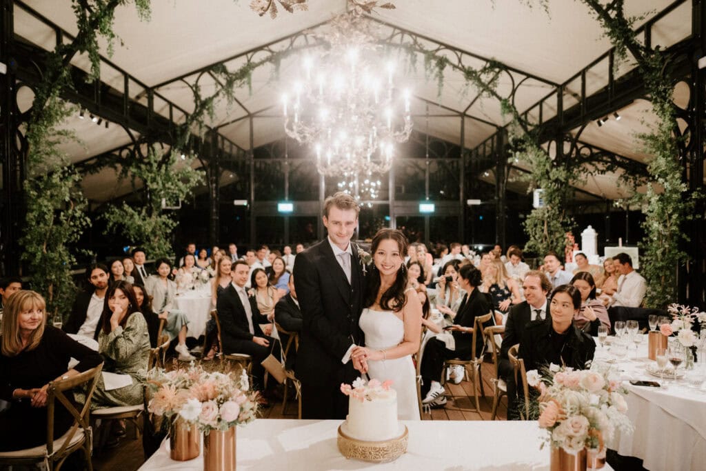 A bride and groom are cutting a wedding cake in an elegant, floral-decorated venue with a large chandelier. Guests are seated at tables around them, watching and smiling.
