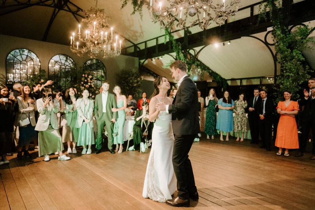 A bride and groom share their first dance on a spacious dance floor, surrounded by guests watching and clapping under chandeliers in an elegantly decorated hall, captured perfectly by the photographer at their stunning Port Lympne wedding.
