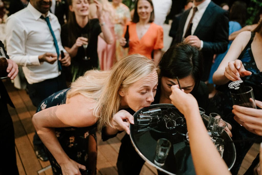 A group of people at a party gather around a tray of drinks, with two women bending down to sip from glasses on the tray. One woman's blonde hair is prominent in the foreground.