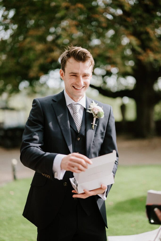 A man in a suit stands outside, smiling while holding an open white box. He wears a boutonniere on his lapel. Trees and grass are visible in the background.