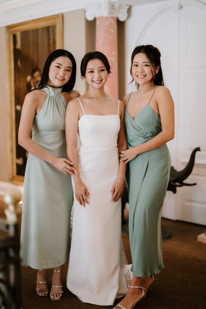 Three women in formal dresses smile and pose for a photo indoors, with the woman in the center wearing a white dress and the other two in green dresses.