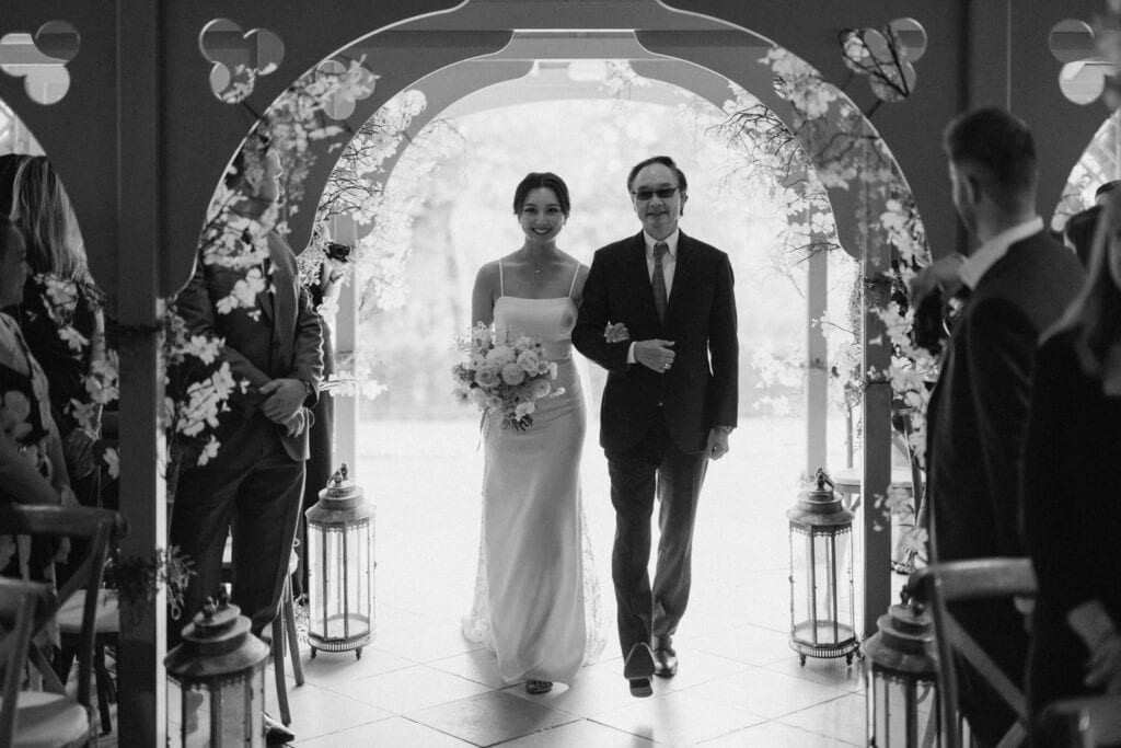 A bride and an older man walk down the aisle in a decorative archway. She holds a bouquet and wears a gown, while he wears a suit and tie. Guests watch as they proceed.