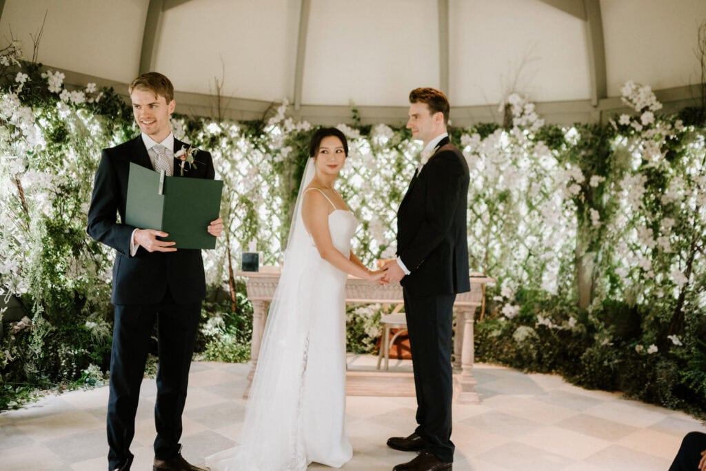 A bride and groom hold hands at the altar during their wedding ceremony, with an officiant standing nearby, holding a green folder. They are in a venue decorated with flowers and greenery.