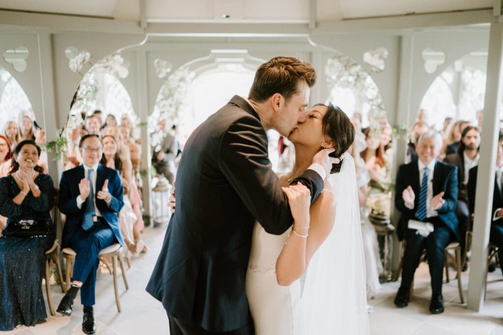A bride and groom share a kiss at their wedding ceremony while guests seated around them applaud. The couple is under a decorated pavilion.