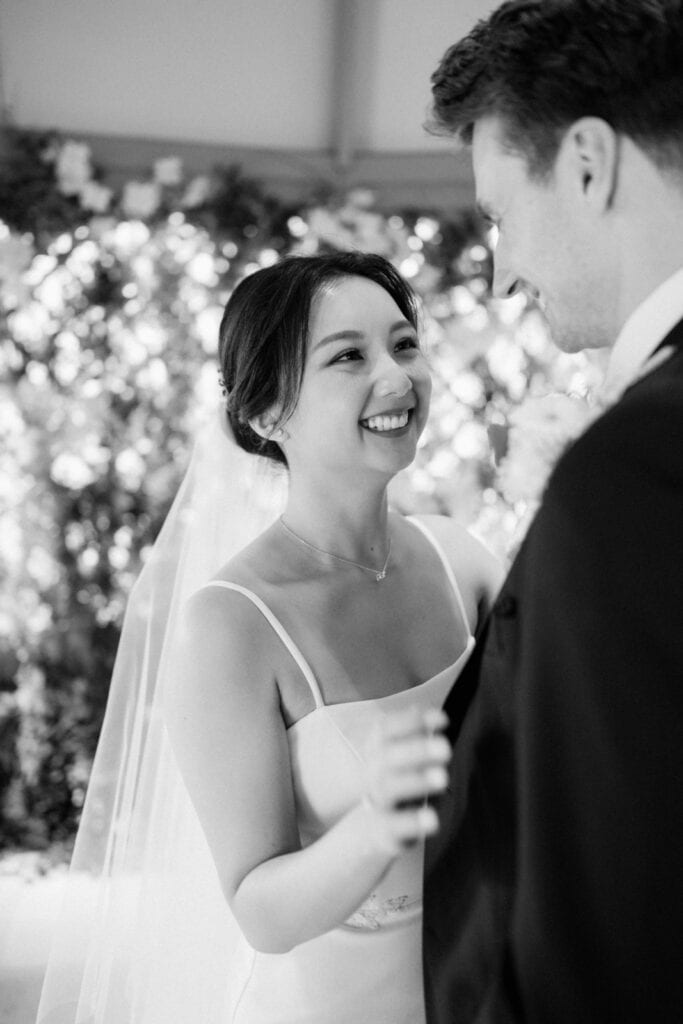 A bride and groom smiling at each other during their wedding ceremony. The bride is wearing a white dress and veil, and the groom is in a suit.