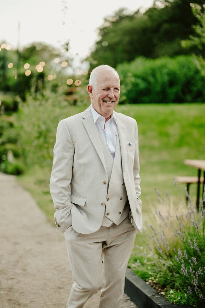 An older man with white hair in a light beige three-piece suit stands outdoors with hands in pockets, smiling. Greenery and string lights are visible in the blurred background.