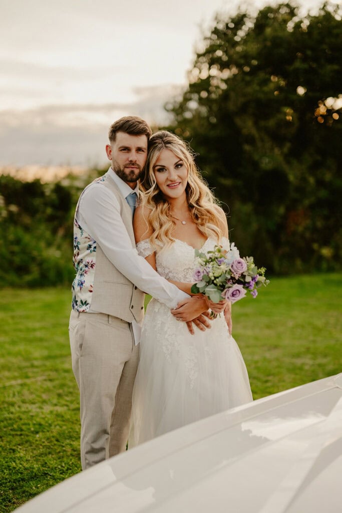 A couple, the man in a beige suit with a floral vest and the woman in a white wedding dress, stand outdoors on a grass field holding a bouquet of flowers.