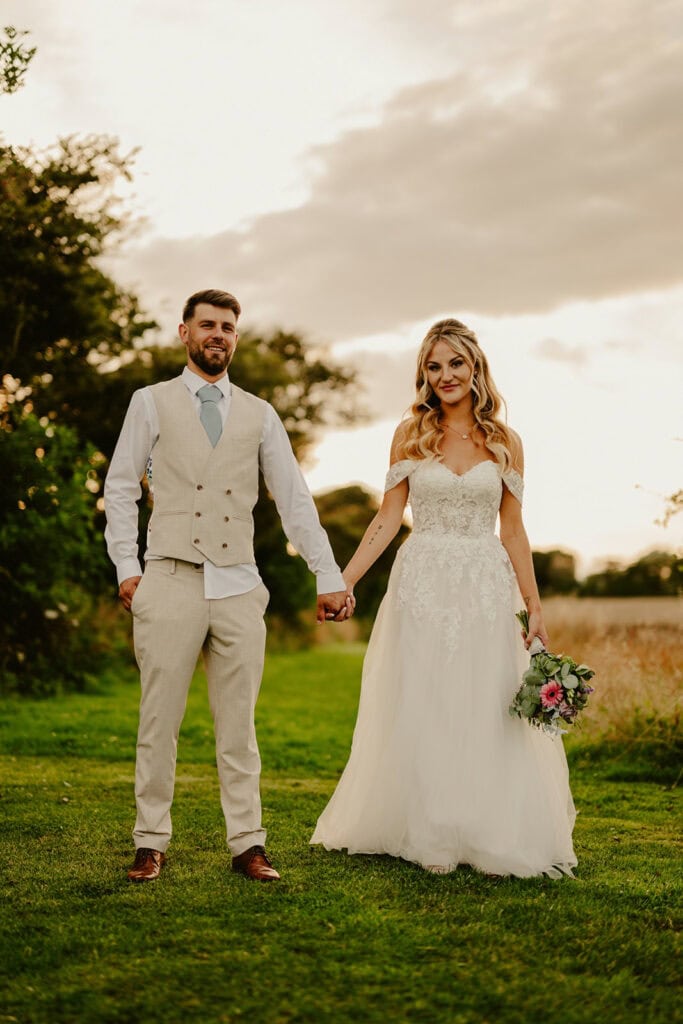 A couple in wedding attire stands hand in hand on a grassy field. The bride holds a bouquet and wears a white gown, while the groom wears a beige suit with a blue tie. Trees and clouds are in the background.