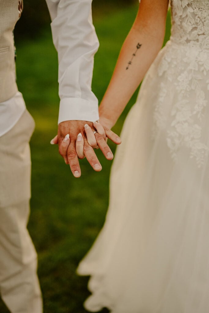 A couple, dressed in wedding attire, holds hands; the man wears a beige suit while the woman wears a white lace wedding dress.