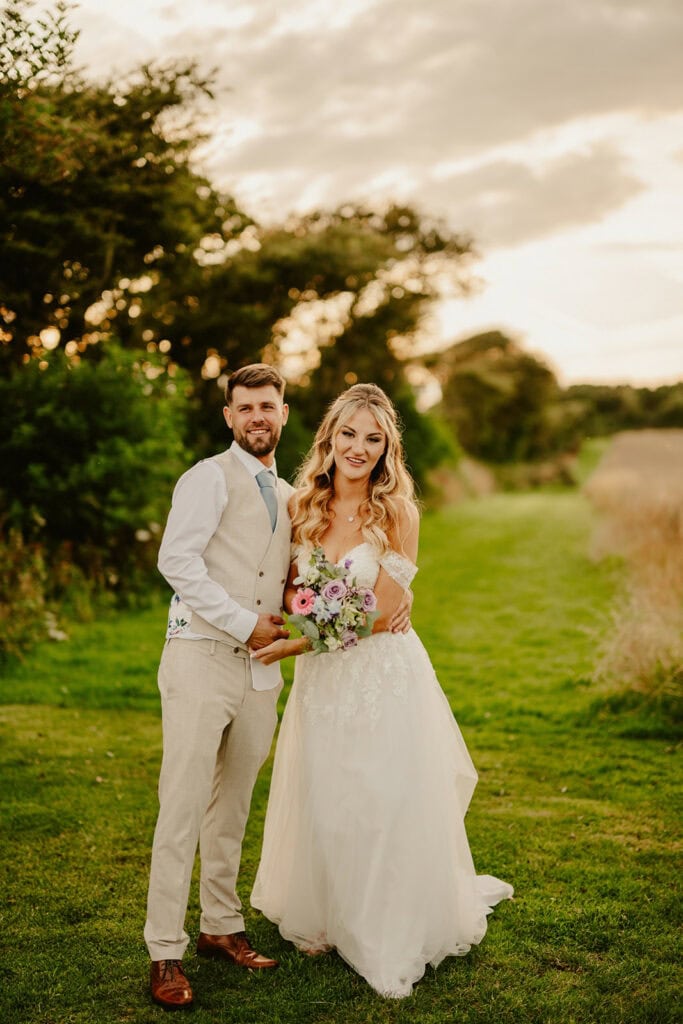A couple in formal wedding attire stands on a grassy path, with the bride holding a bouquet of flowers. Trees and a cloudy sky are in the background.