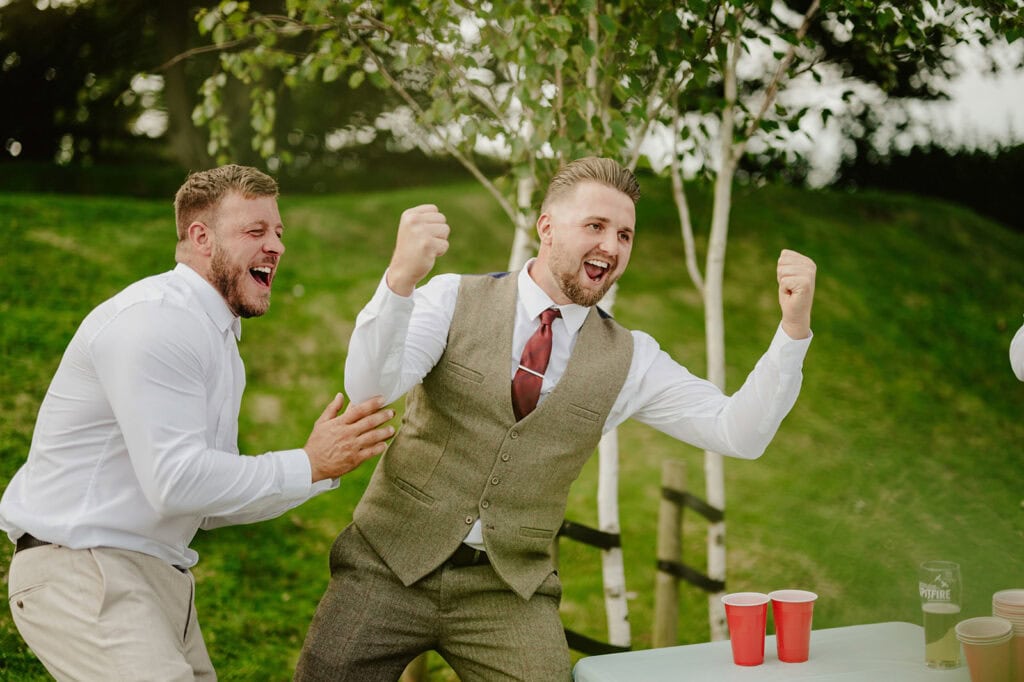 Two men in white shirts, one in a vest and tie, cheer enthusiastically by a table with red cups in an outdoor setting with green trees in the background.