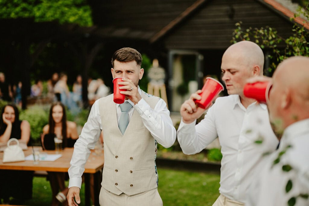Two men in formal attire drink from red cups at an outdoor gathering, with a group of people and tables in the background.