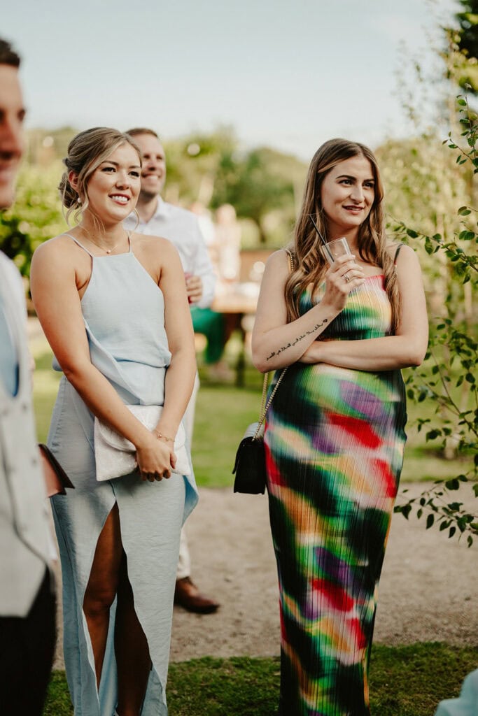 Two women, one in a light blue dress and the other in a colorful dress, stand outdoors at an event. Both are smiling, with the second woman holding a drink. Other people and greenery are in the background.