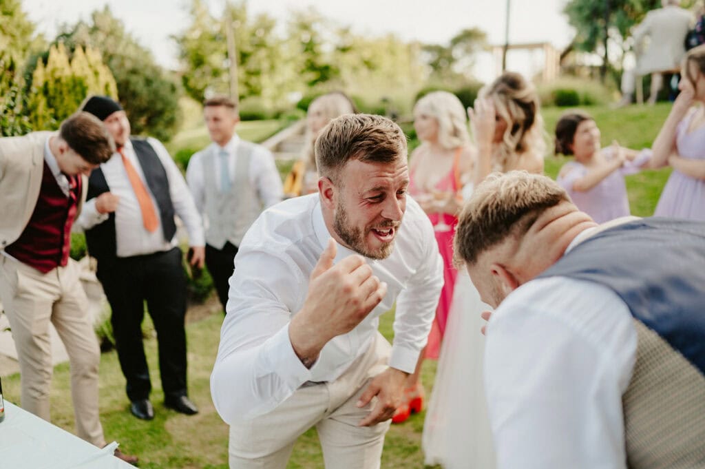A man in a white shirt and light pants enthusiastically gestures while talking to another man in a vest. Several people are seen in the background at an outdoor event.