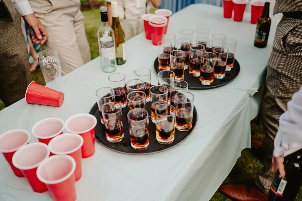 A table is set with trays of drinks in glasses, red plastic cups, and several bottles of wine and beer. People are standing around the table.