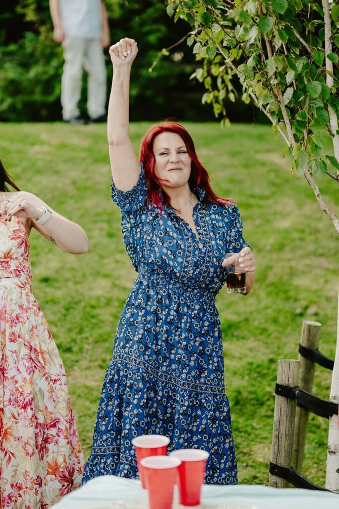 A woman in a blue floral dress raises her fist in the air while holding a drink in the other hand, standing outside near a table with red cups and a tree in the background.