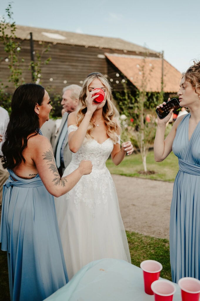 Three women in formal dresses drinking from cups at an outdoor event. The woman in the center is in a bridal gown, while the others are in light blue dresses. A building and trees are in the background.