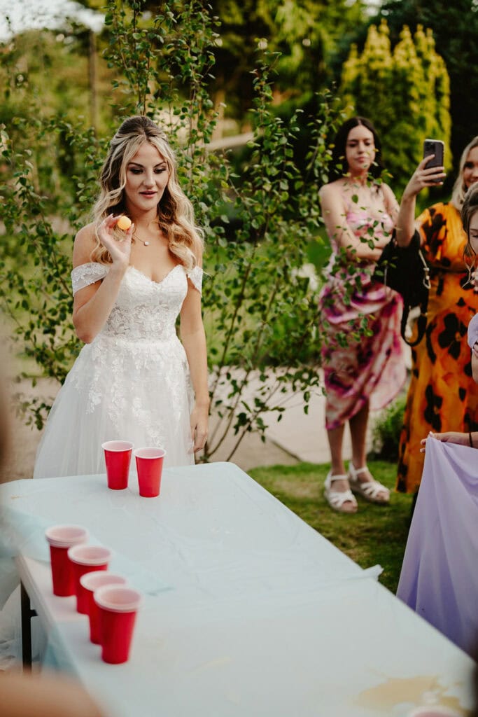A woman in a white wedding dress plays a drinking game with red cups on a table outdoors. People surround her, and some are taking pictures.