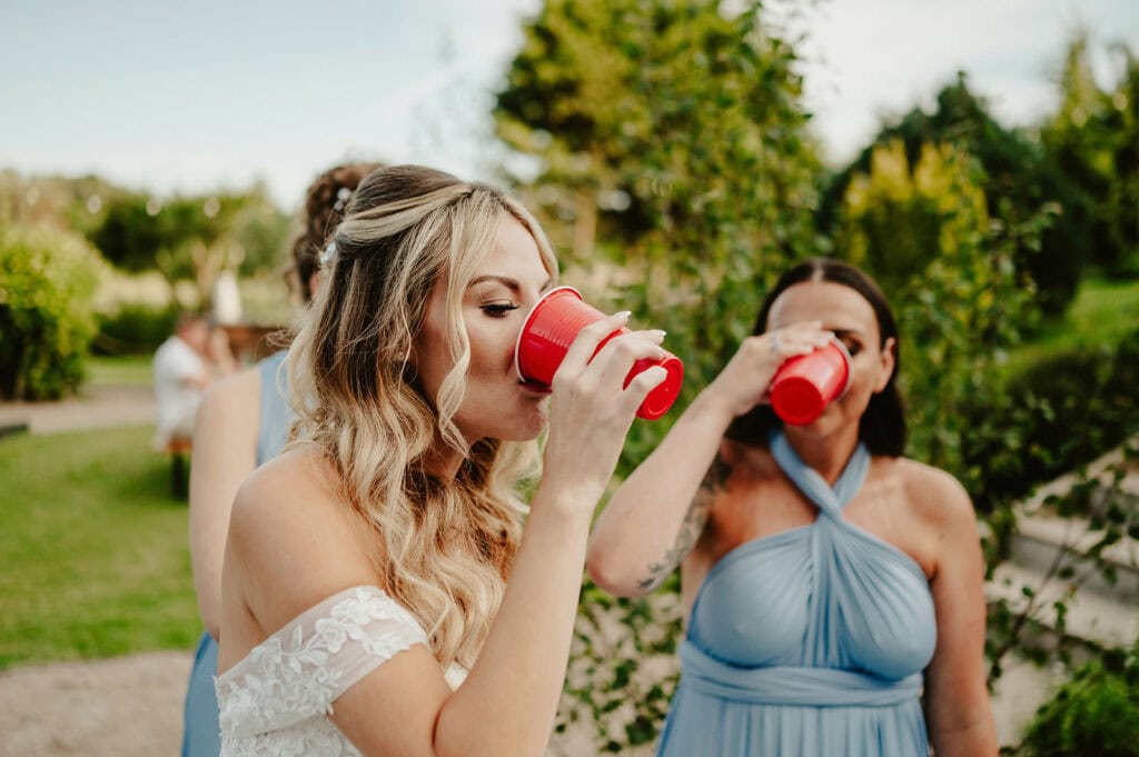 A woman in a white dress and another in a blue dress drink from red cups outdoors, with trees and grass in the background.
