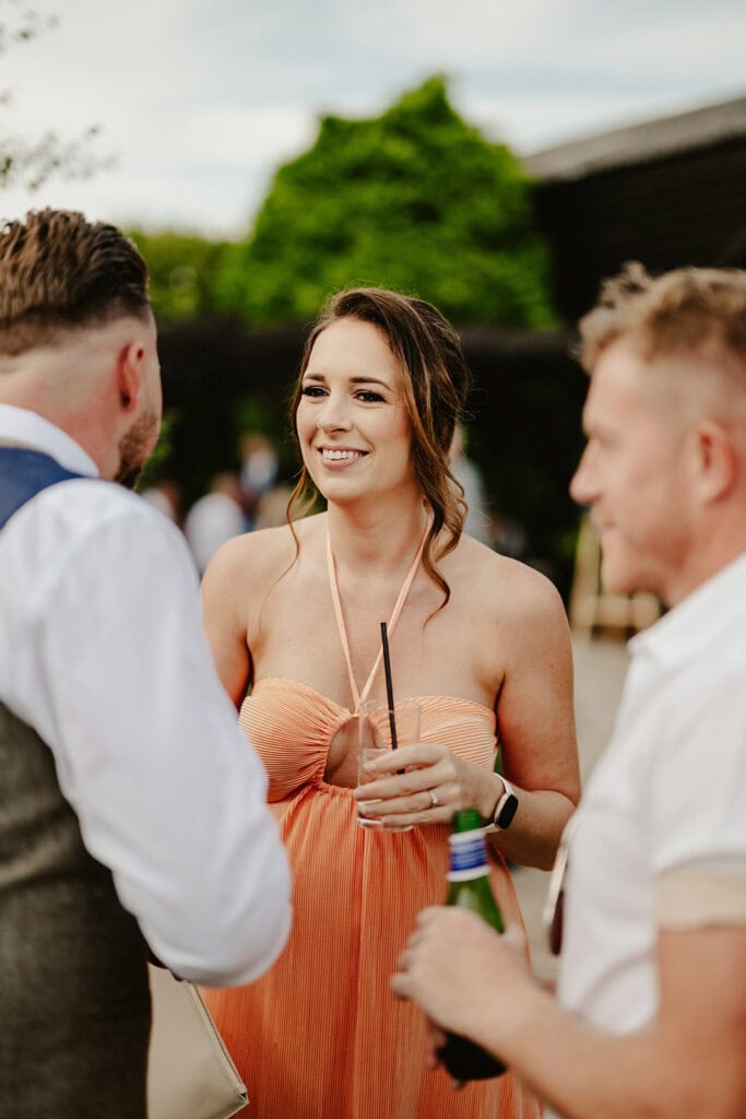 A woman in an orange dress holding a drink stands outdoors, smiling and conversing with two men, one in a white shirt and the other in a vest and tie.