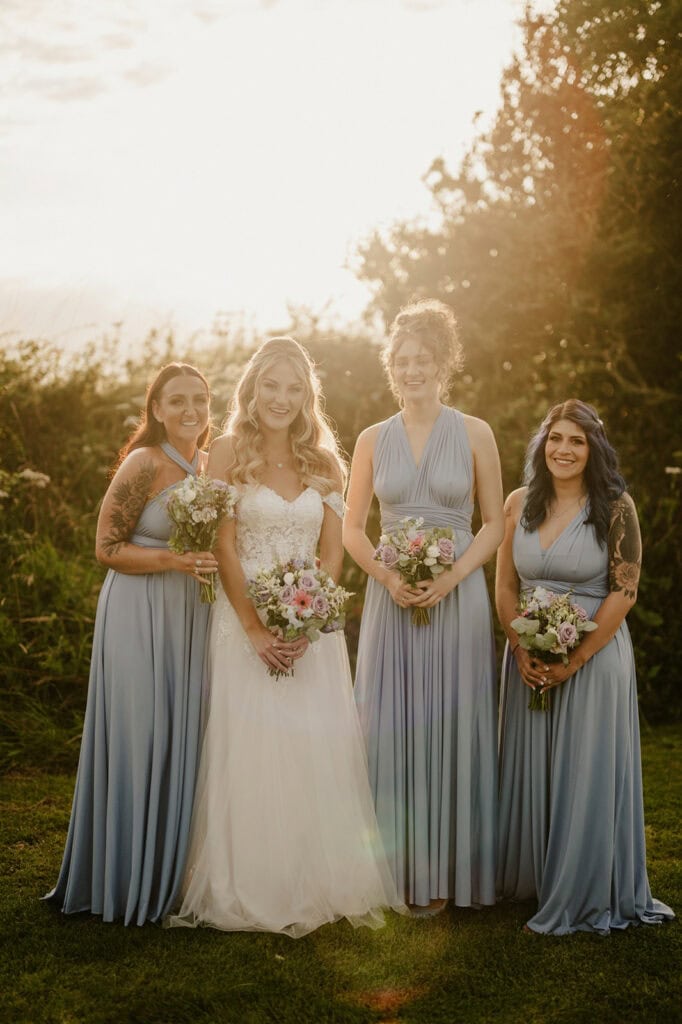 Four women in formal dresses stand outdoors, holding floral bouquets. One wears a white wedding gown, while the other three wear light blue bridesmaid dresses. Trees and sunlight are in the background.