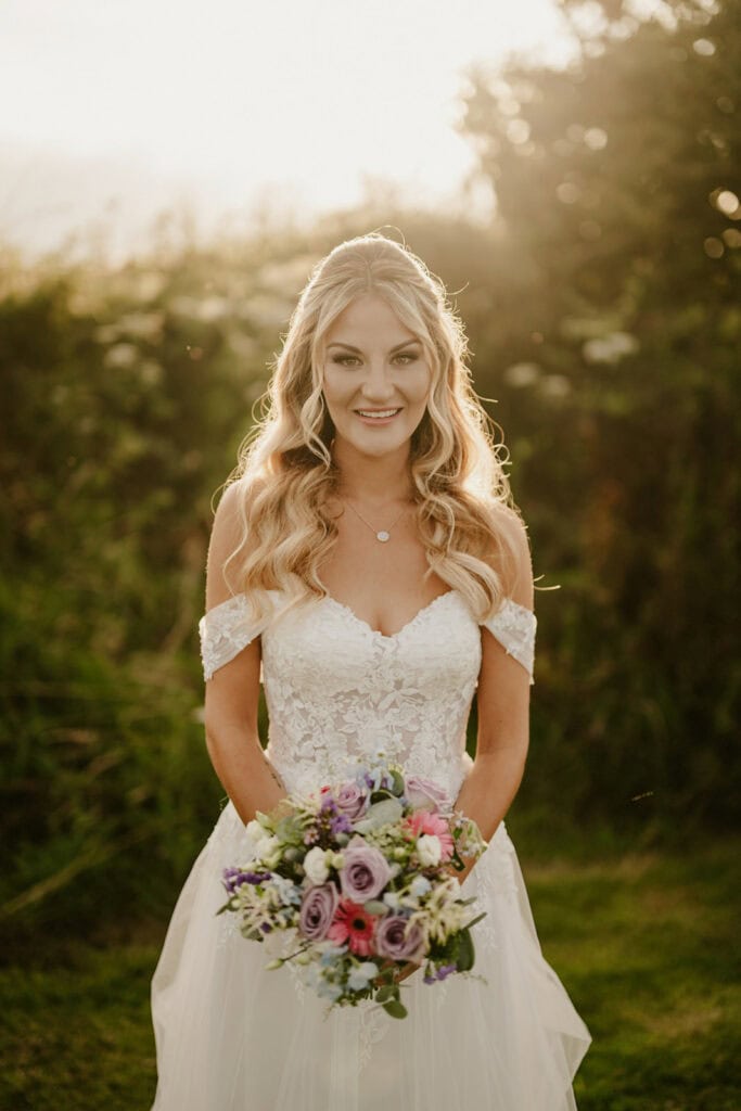 A bride in a white off-shoulder lace wedding dress holds a colorful bouquet while standing outdoors with greenery and sunlight in the background.