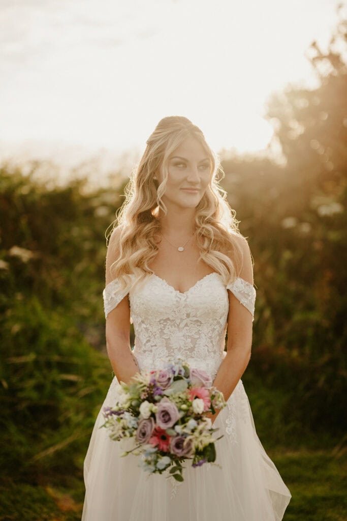 A woman in a wedding dress with lace details holds a bouquet of flowers against a backdrop of greenery and sunlight.