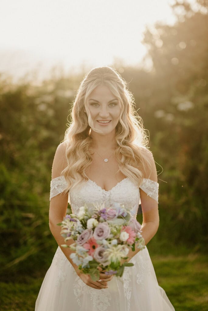 A bride with long blonde hair stands outdoors holding a bouquet of flowers. She is wearing a white off-the-shoulder wedding dress, and greenery is visible in the background.