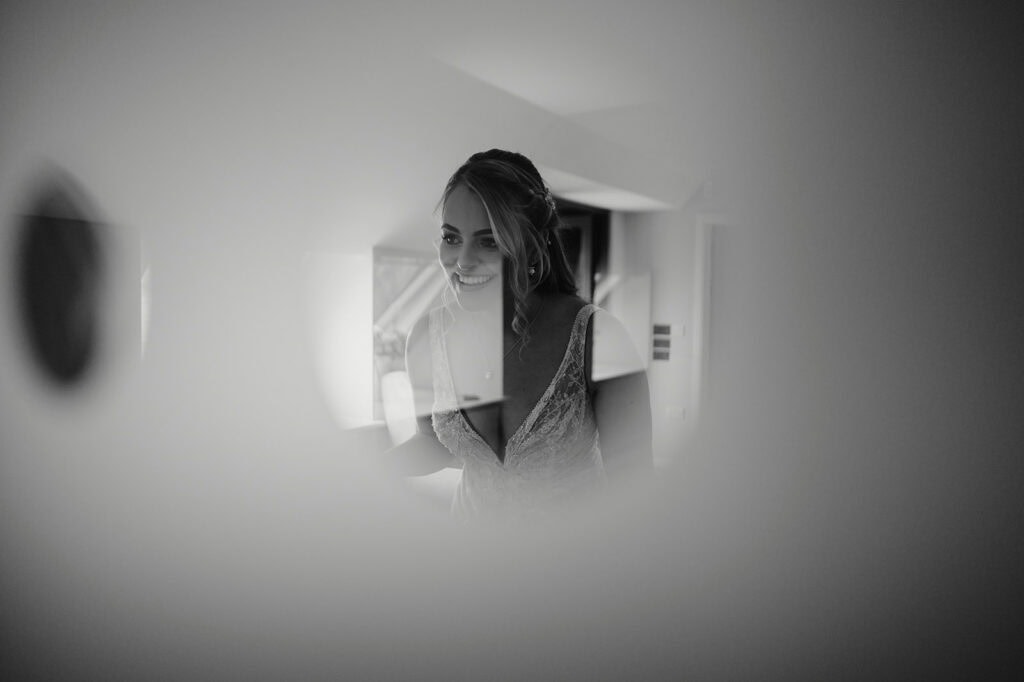 A woman in a wedding dress smiles as seen through a circular opening, framed in soft focus. Black and white image.