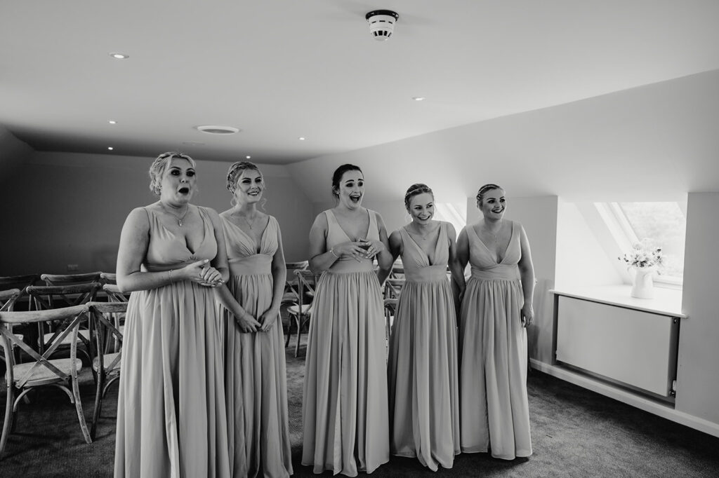 Five women in matching sleeveless dresses stand in a line, smiling and looking in one direction in a brightly lit room with chairs and a window.