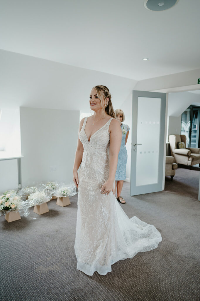 A woman in a white lace wedding dress stands in a room with bouquets of flowers on the floor and another woman in a light blue dress behind her.