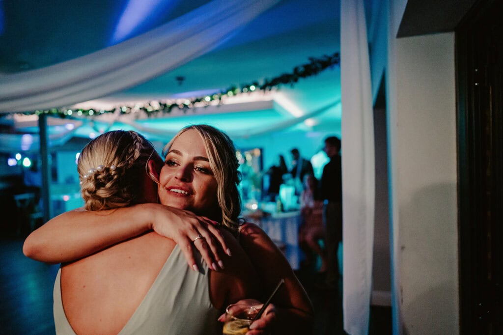 Two people embracing in a warmly lit room, with one holding a drink. The background shows other people and tables under soft blue lighting.