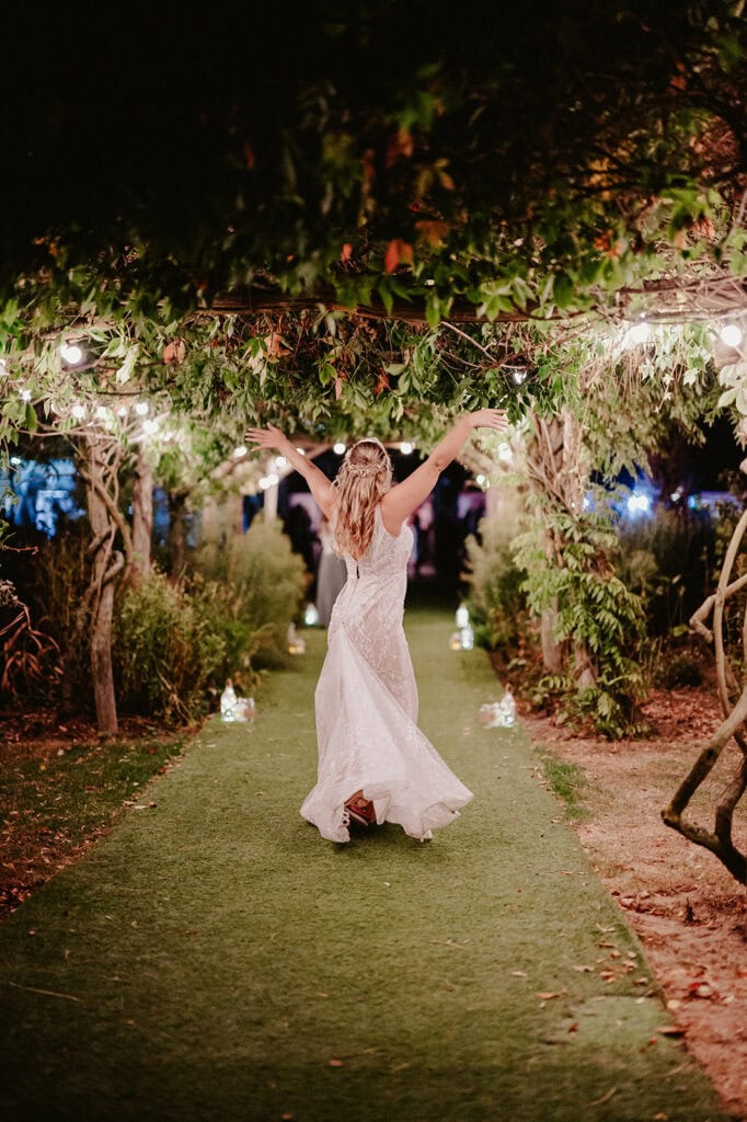 A woman in a white wedding dress walks down an outdoor, tree-covered path decorated with lights at night, with her arms raised in celebration.