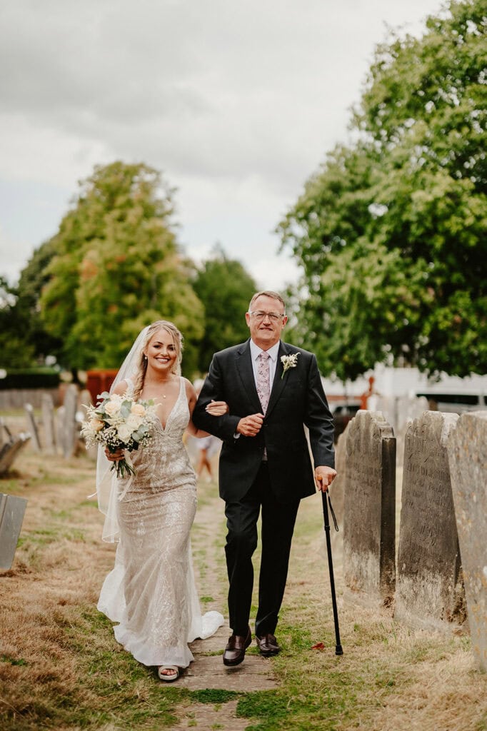 A bride in a white wedding dress walks arm-in-arm with an older man in a black suit holding a cane, outdoors on a grassy path lined with gravestones.