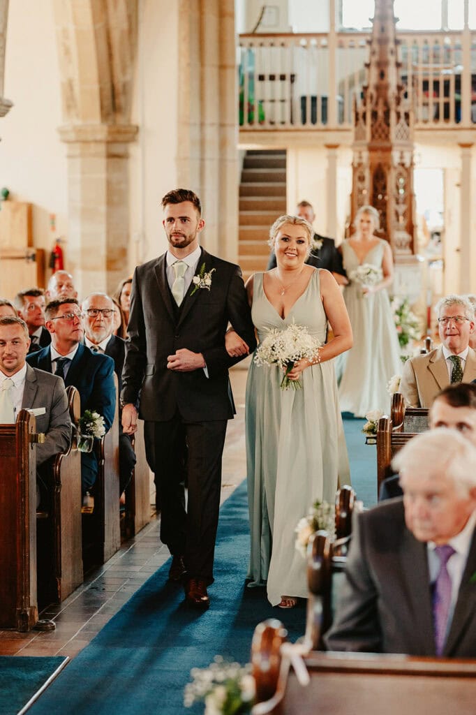 A bridesmaid and groomsman walk down the aisle of a church during a wedding ceremony, surrounded by seated guests.
