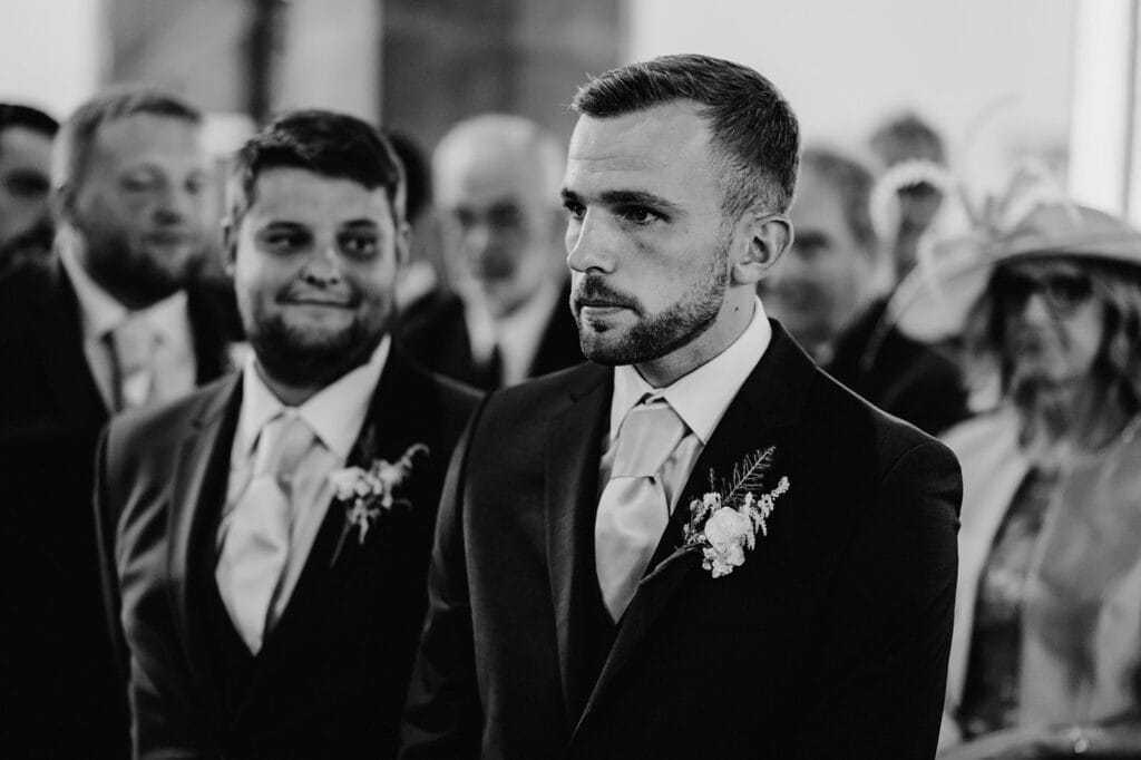 A groom, neatly dressed in a suit with a floral boutonniere, stands beside a groomsman in a similar outfit, surrounded by people in formal attire in a black and white indoor setting.
