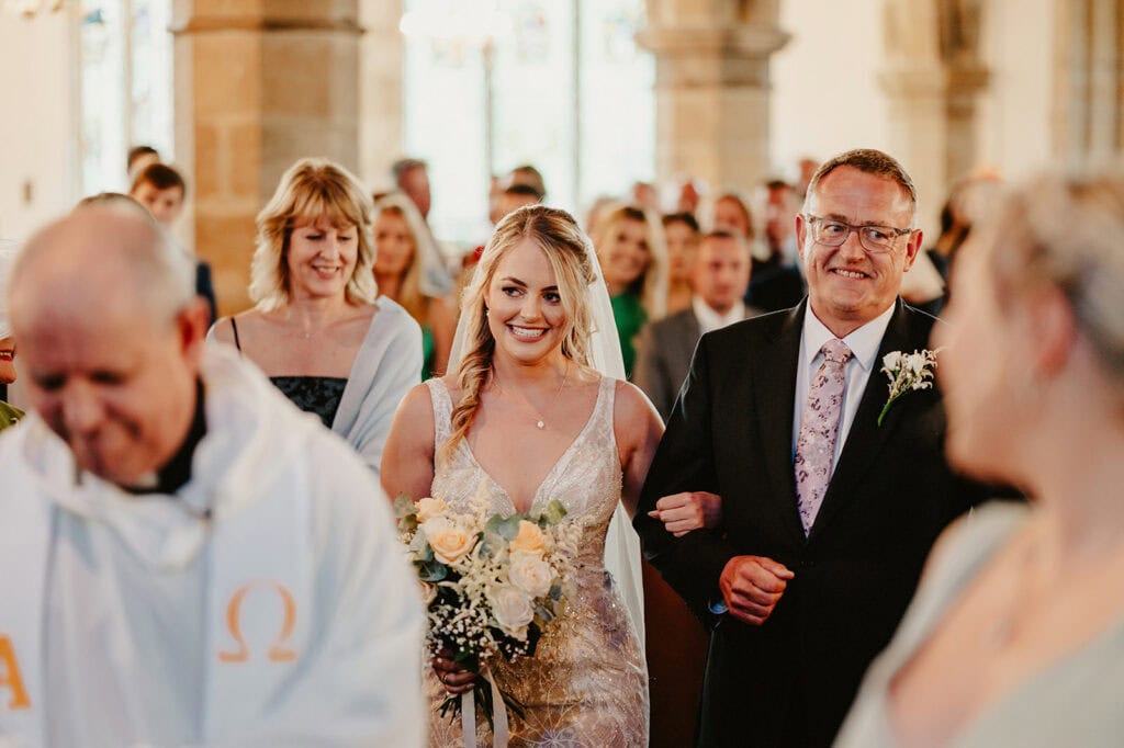 A bride, holding a bouquet, walks down the aisle arm-in-arm with an older man, followed by a woman. A priest stands in the foreground in a brightly lit church filled with guests.