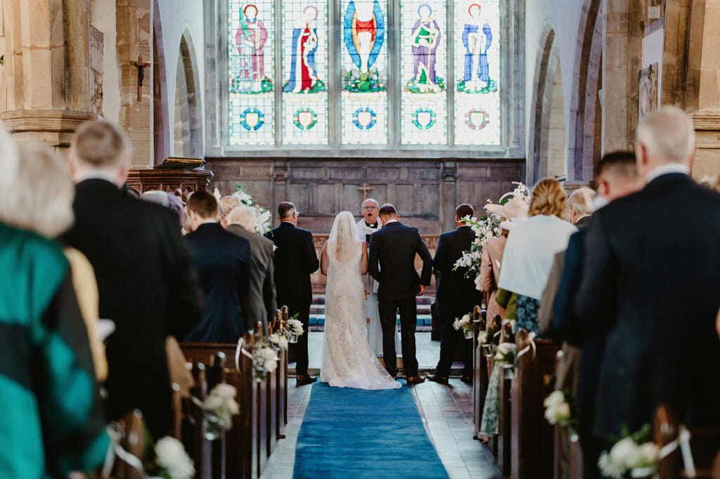 A wedding ceremony taking place in a church with the bride and groom standing at the altar. Attendees are seated on either side of the aisle, and stained glass windows are visible in the background.