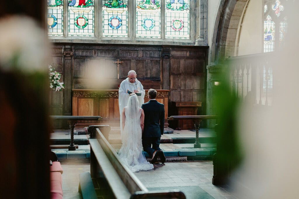 A bride and groom kneel before a clergy member in a church with stained glass windows and wooden paneling.