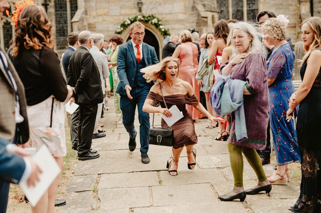 A woman in a brown dress is tripping and falling forward on a stone pathway amidst a crowd of people dressed formally. Some onlookers appear surprised while others continue to converse.