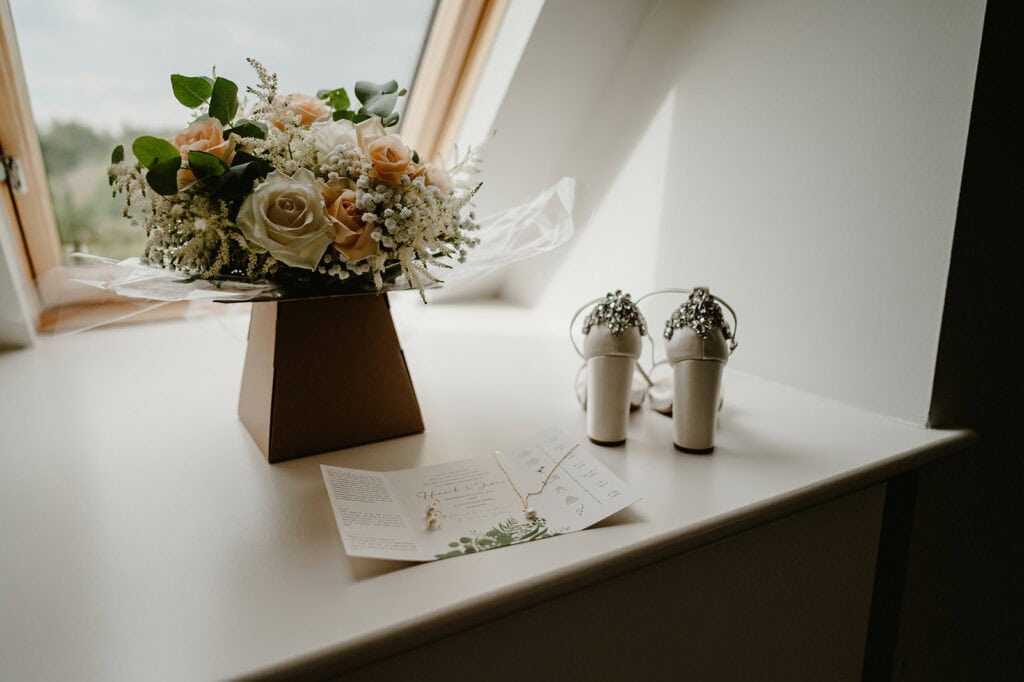 A bouquet of flowers, wedding invitation, necklace, and pair of white high-heeled shoes are placed on a windowsill.