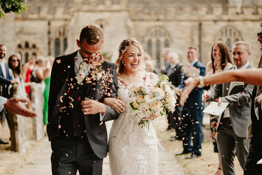 Newlyweds walk down an outdoor aisle, smiling and holding hands, as guests throw confetti. The bride holds a bouquet and wears a white dress, while the groom is in a black suit.
