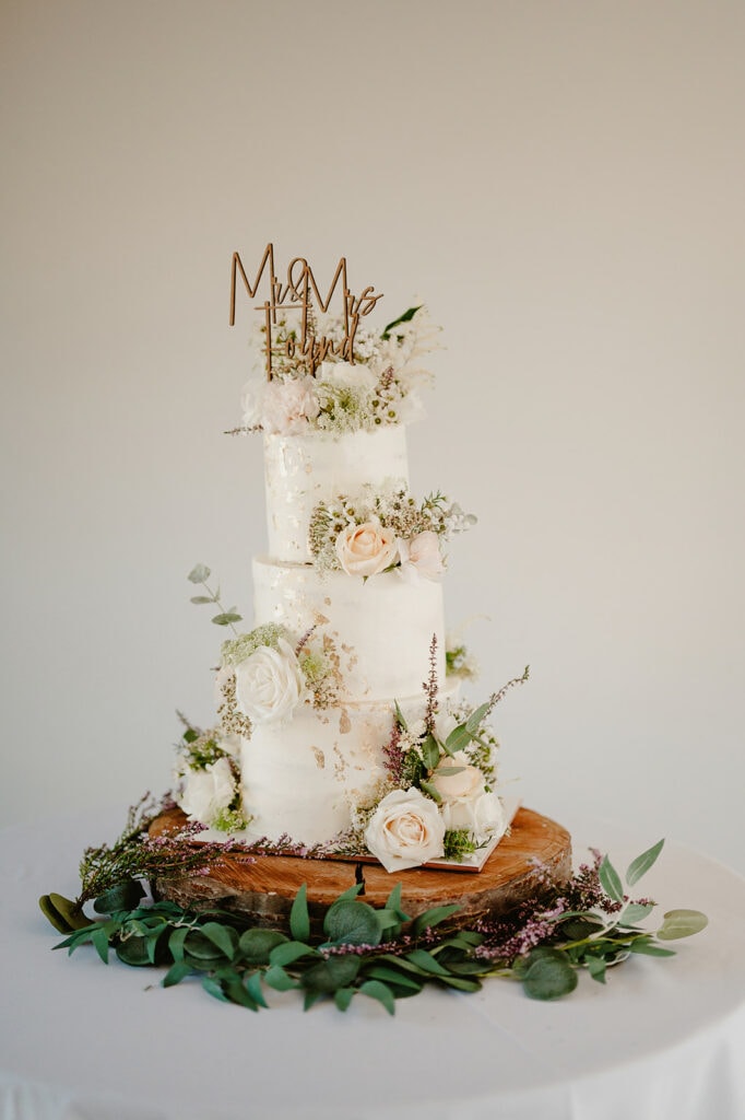 Three-tier white wedding cake decorated with white and blush roses, greenery, and gold accents. A gold "Mr & Mrs" topper sits on the highest tier. Cake is displayed on a wooden base.