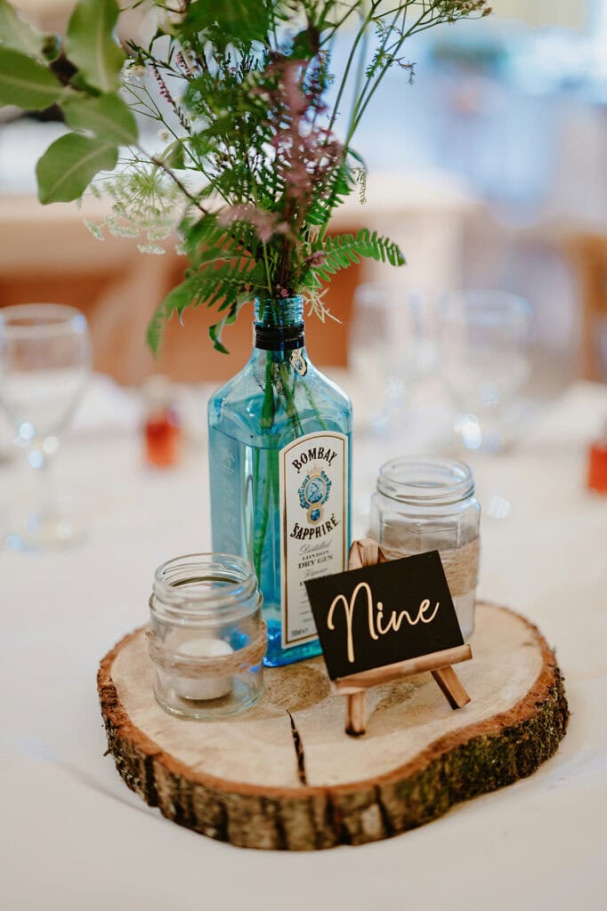A centerpiece with a blue gin bottle used as a vase holding greenery, two mason jars, and a small sign reading "Nine" on a wooden slice.