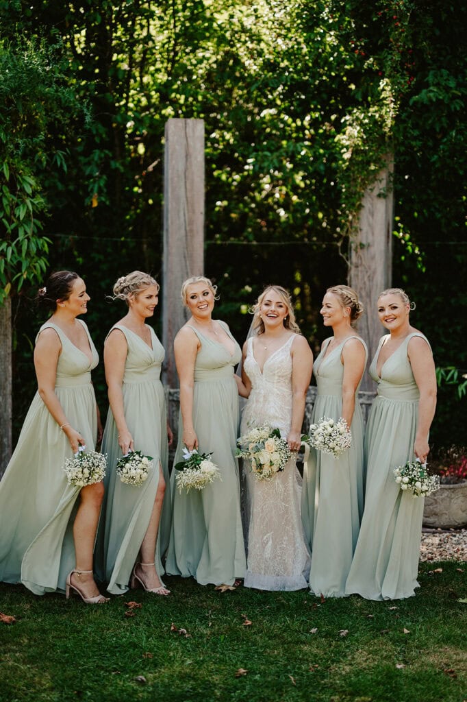A bride in a white dress stands with five bridesmaids in light green dresses, holding bouquets, posing outdoors in a lush garden setting.