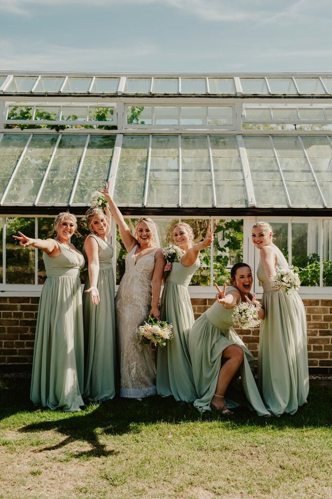 A bride and four bridesmaids in light green dresses pose happily in front of a greenhouse on a sunny day, holding bouquets and smiling.