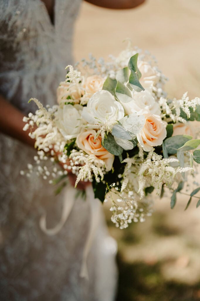 A person in a lace dress holding a bouquet of white and peach roses, eucalyptus leaves, and white accent flowers.