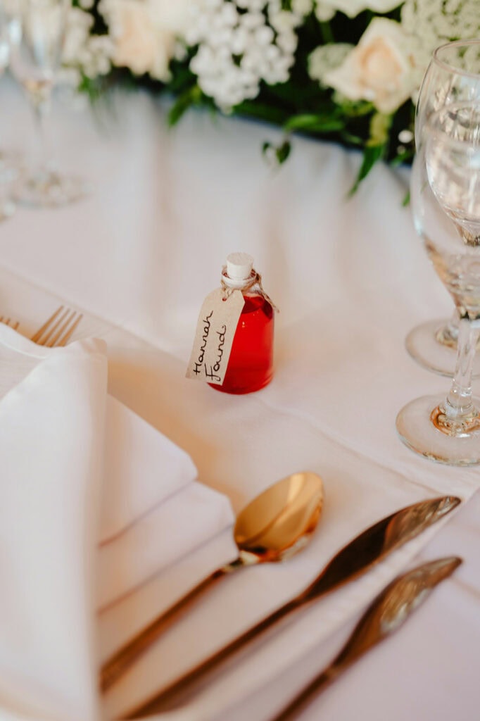 Table setting with white napkin, gold cutlery, wine glasses, and a small bottle with red liquid labeled "Friends Forever." Floral arrangement in the background.