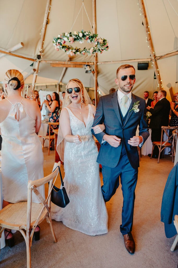 A bride and groom wearing sunglasses walk arm-in-arm in a wedding tent. The bride wears a white gown, and the groom is in a blue suit. Guests seated at tables are seen in the background.