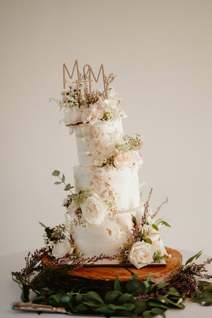 A three-tiered wedding cake decorated with white flowers, greenery, and gold accents. The cake topper reads "Mr & Mrs Howard." The cake is set on a wooden stand against a plain background.